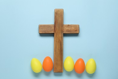 Wooden cross and painted Easter eggs on light blue background, flat lay