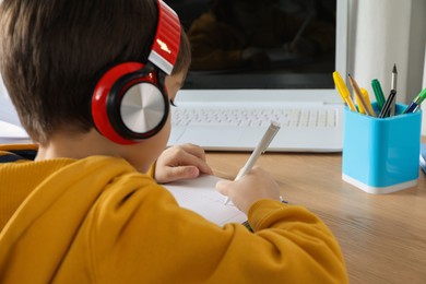 Photo of Cute little boy with headphones and modern laptop studying online at home. E-learning