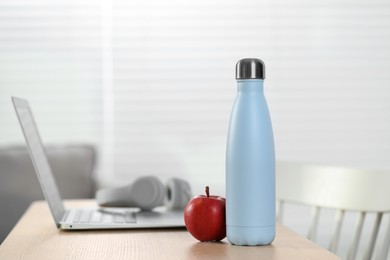 Photo of Stylish thermo bottle, apple and laptop on wooden table at home. Space for text