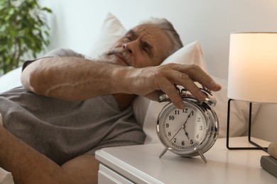 Photo of Sleepy senior man turning off alarm clock in bedroom, focus on hand