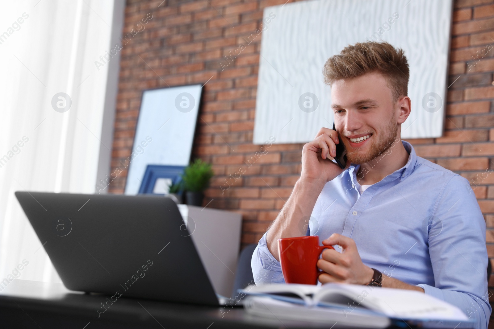 Photo of Young man talking on phone while using laptop at table indoors