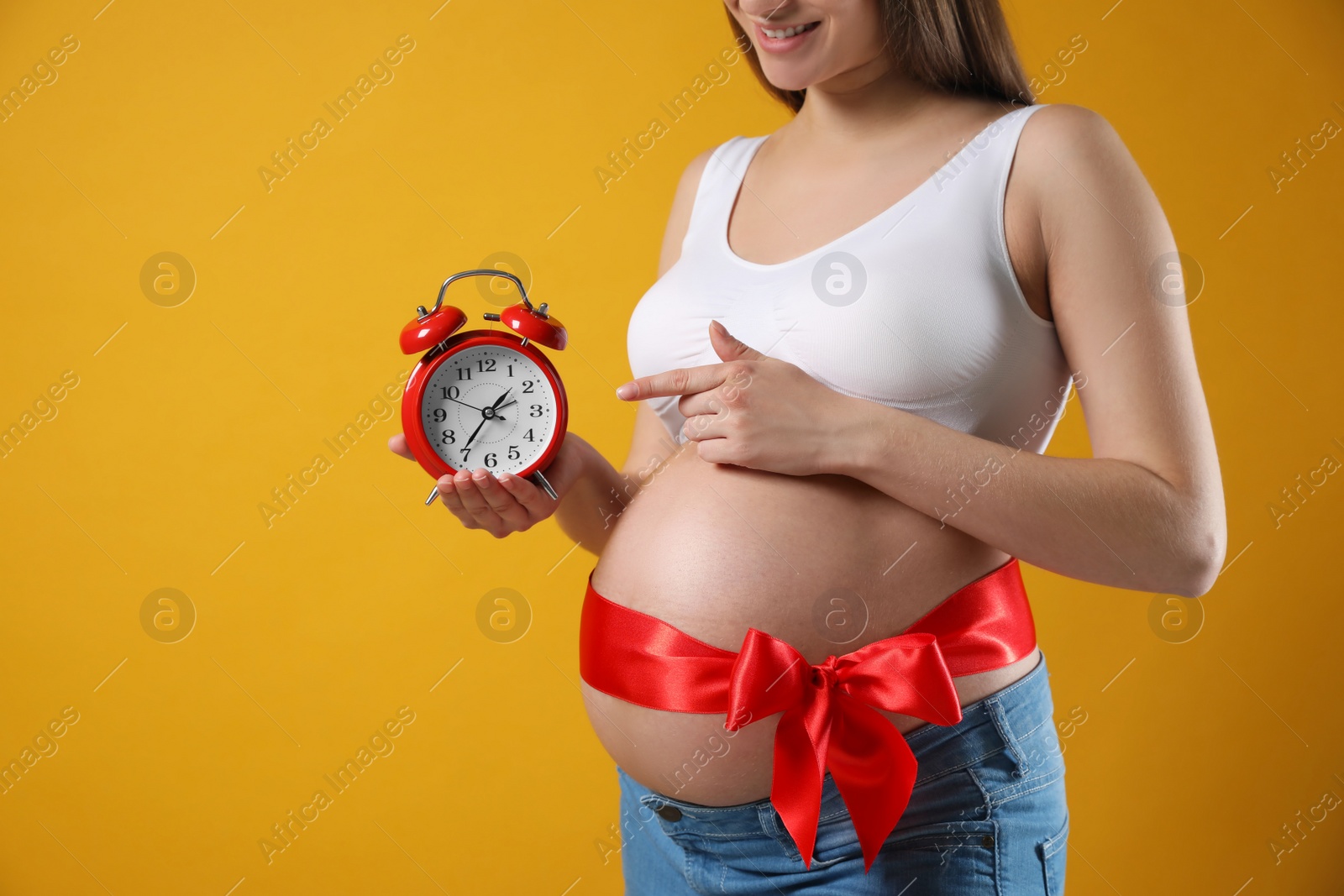 Photo of Young pregnant woman with alarm clock and red bow on yellow background, closeup. Time to give birth