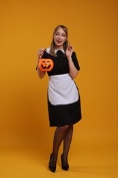 Photo of Happy woman in scary maid costume with pumpkin bucket on orange background. Halloween celebration