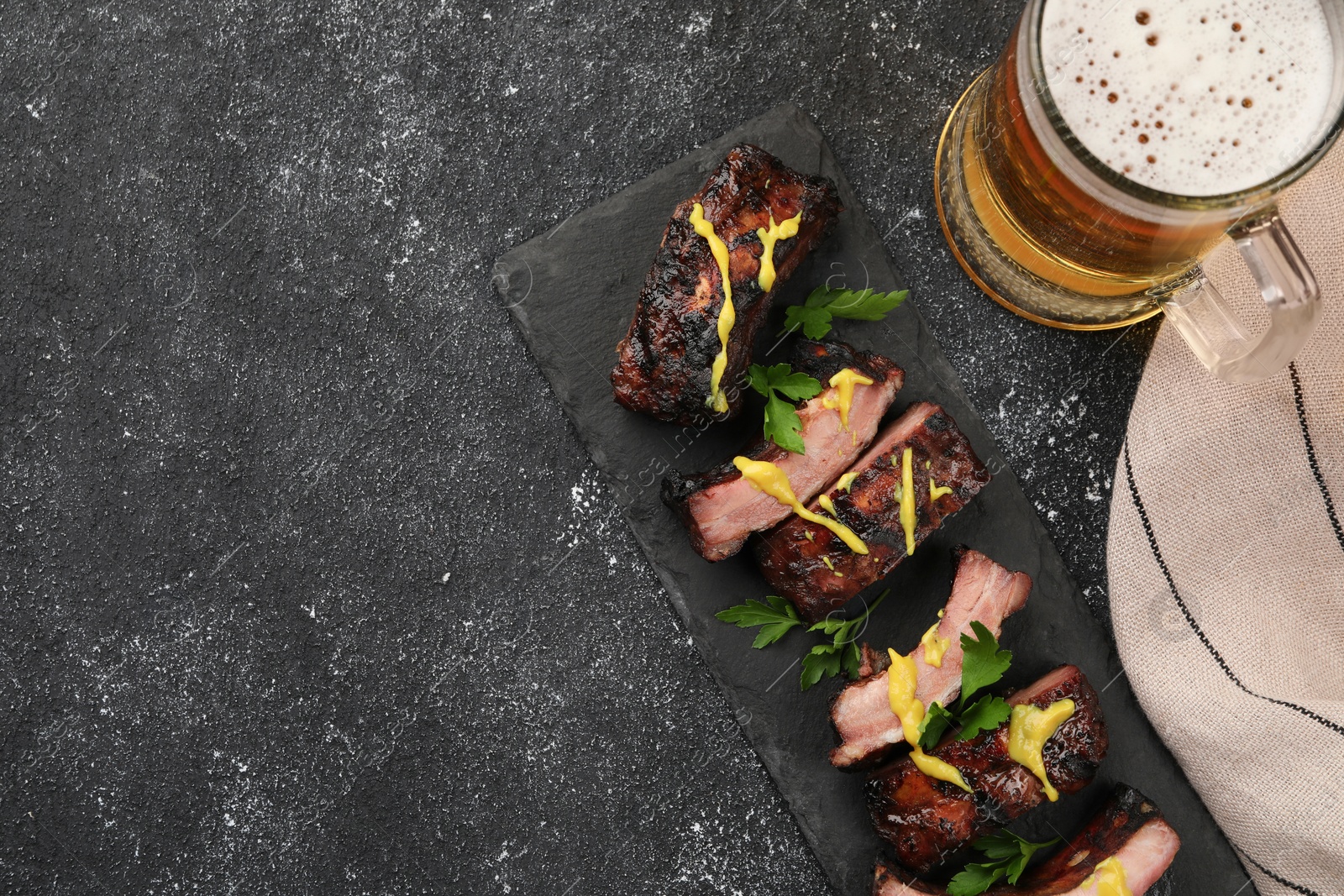 Photo of Mug with beer and delicious grilled ribs on dark grey textured table, flat lay. Space for text