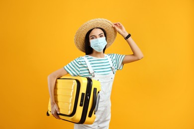 Female tourist in medical mask with suitcase on yellow background. Travelling during coronavirus pandemic