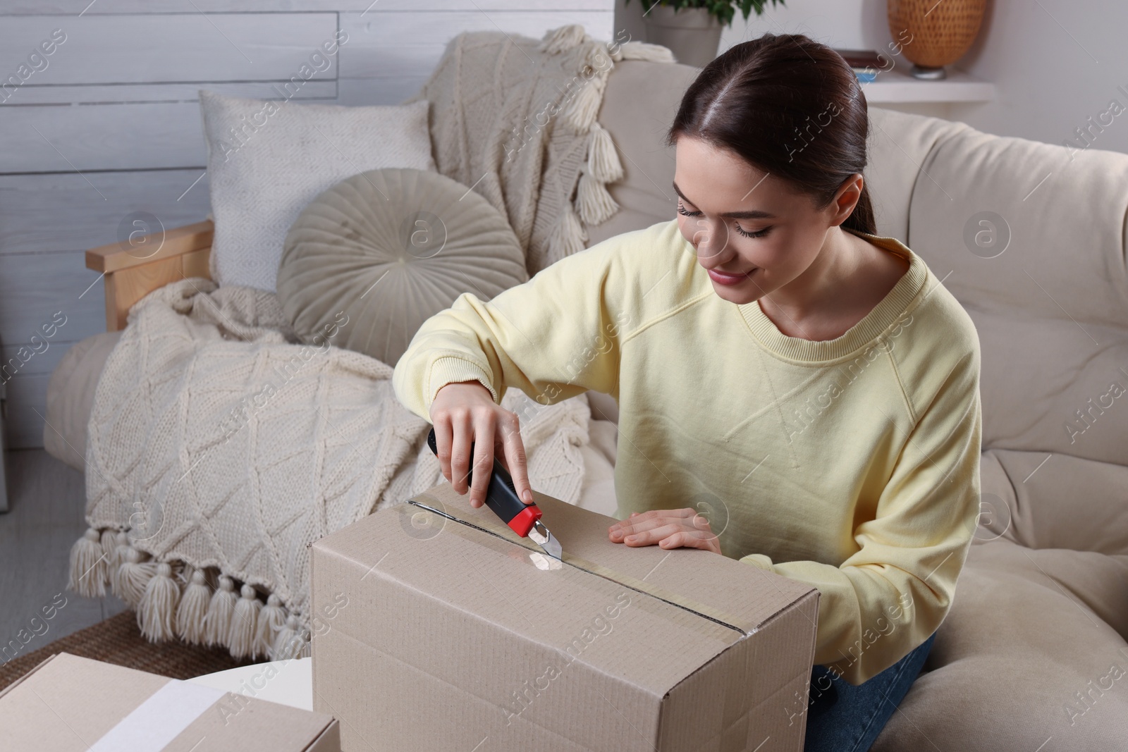 Photo of Young woman using utility knife to open parcel at home