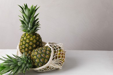 Photo of Net bag with delicious ripe pineapples on white table near light grey wall. Space for text