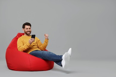 Photo of Happy man with smartphone sitting on bean bag against grey background. Space for text