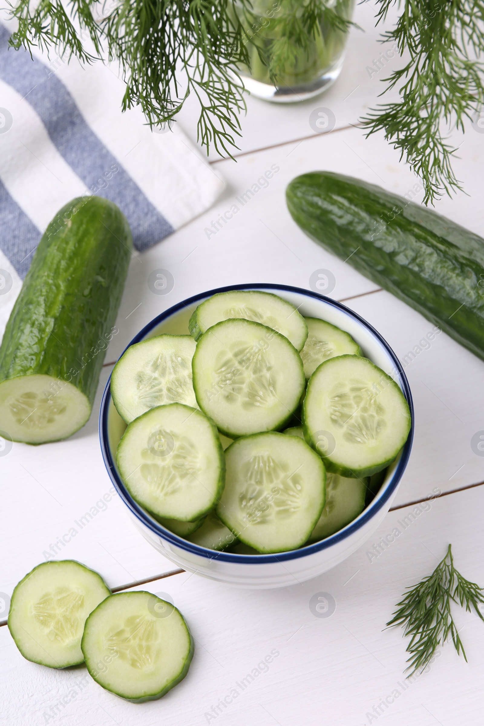 Photo of Cut cucumber in bowl, fresh vegetables and dill on white wooden table, above view