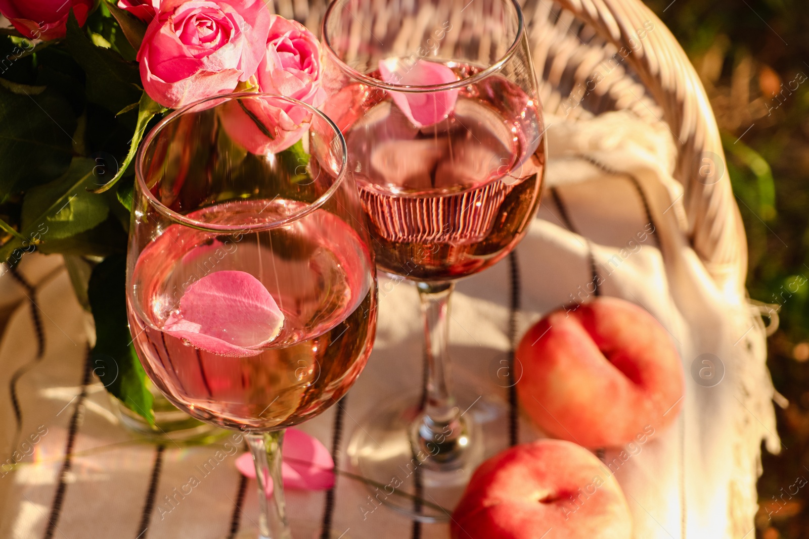 Photo of Glasses of delicious rose wine with petals, peaches and flowers on white picnic blanket outside