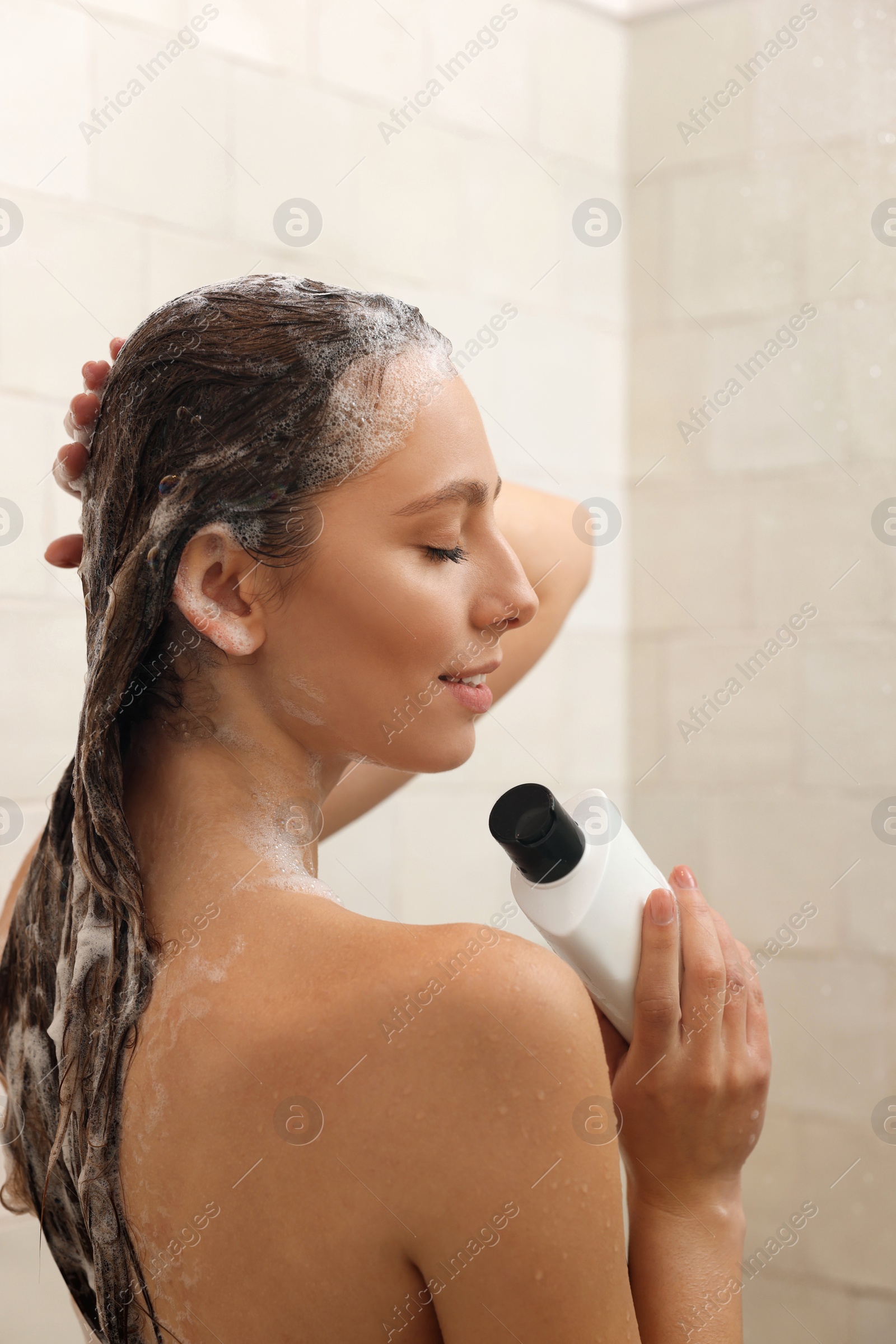 Photo of Young woman with bottle of shampoo in shower at home. Washing hair