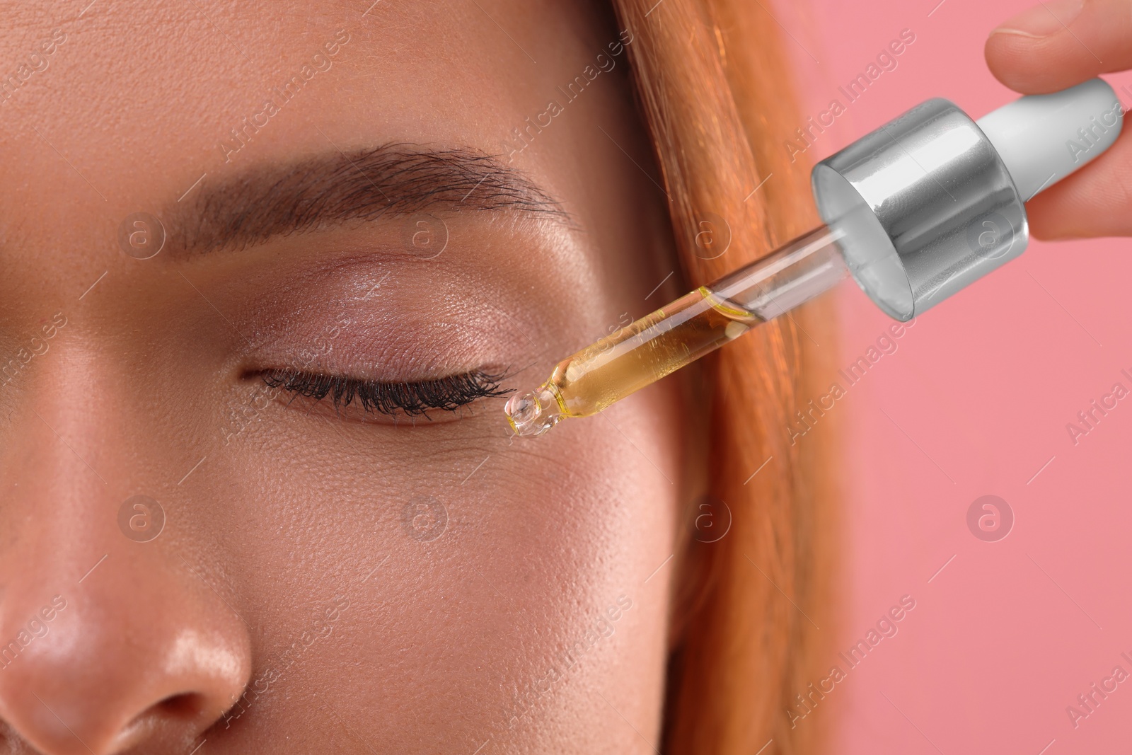 Photo of Beautiful young woman applying cosmetic serum onto her face on pink background, closeup