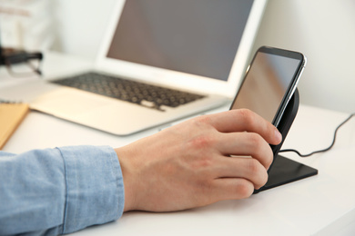 Photo of Man putting mobile phone onto wireless charger at white table, closeup. Modern workplace accessory