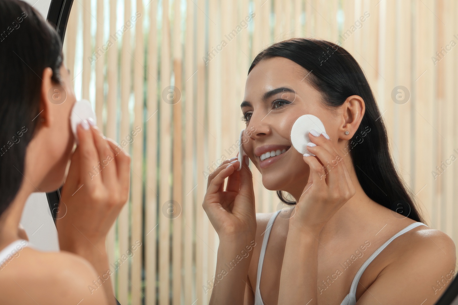 Photo of Young woman using cotton pads with micellar water near mirror indoors