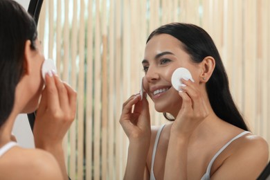 Young woman using cotton pads with micellar water near mirror indoors