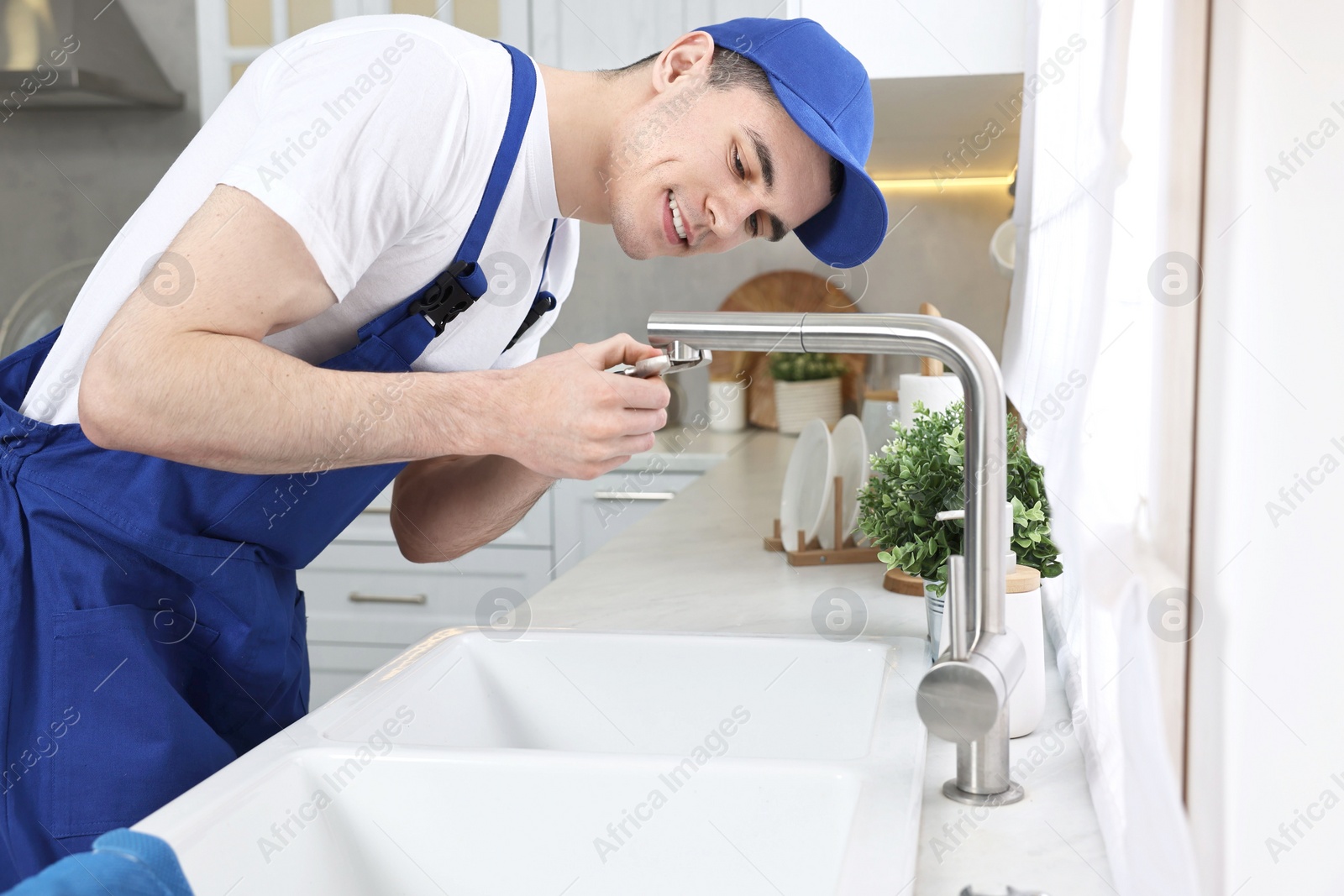 Photo of Smiling plumber repairing faucet with spanner in kitchen