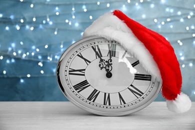 Clock with Santa hat on table against blurred lights. Christmas countdown