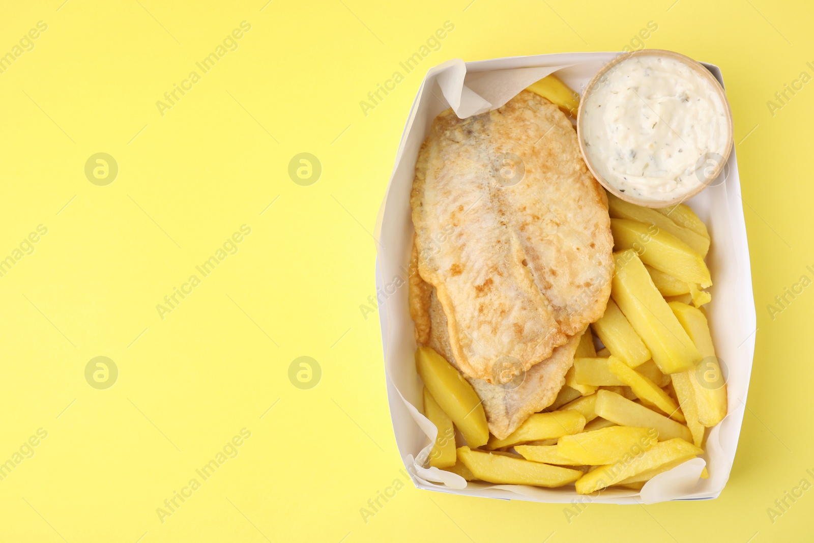 Photo of Delicious fish and chips with tasty sauce in paper box on yellow table, top view. Space for text