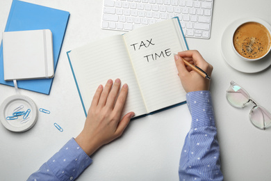 Image of Woman writing words TAX TIME in notebook at white table, top view