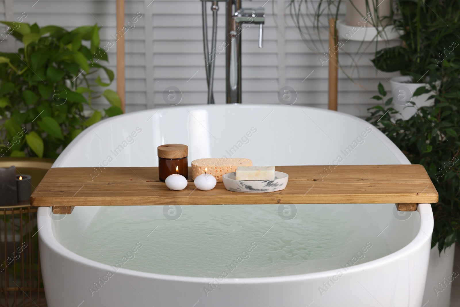 Photo of Wooden board with spa products and burning candles on bath tub in bathroom