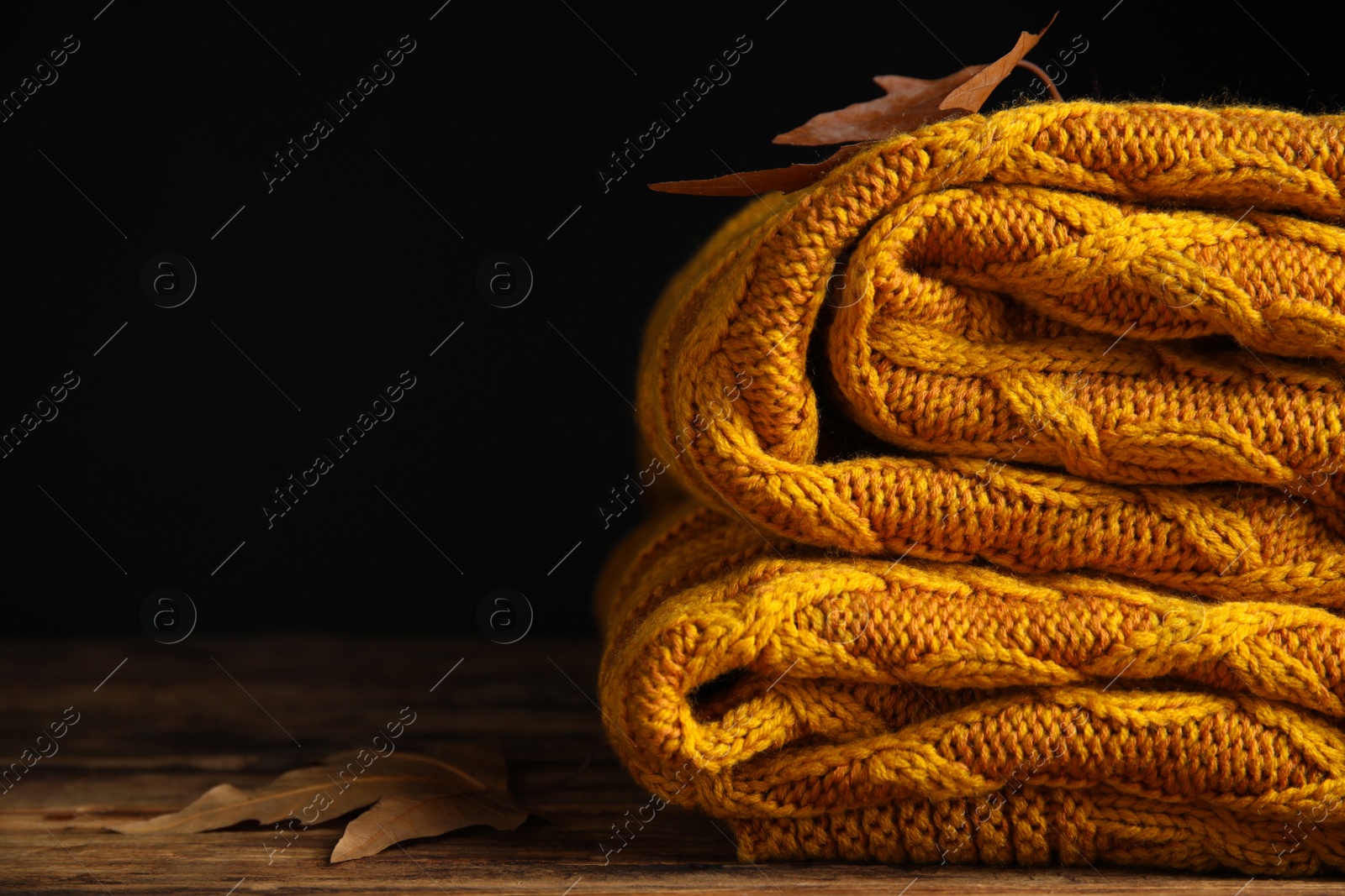 Photo of Orange knitted plaid with dry leaves on wooden table, closeup. Space for text