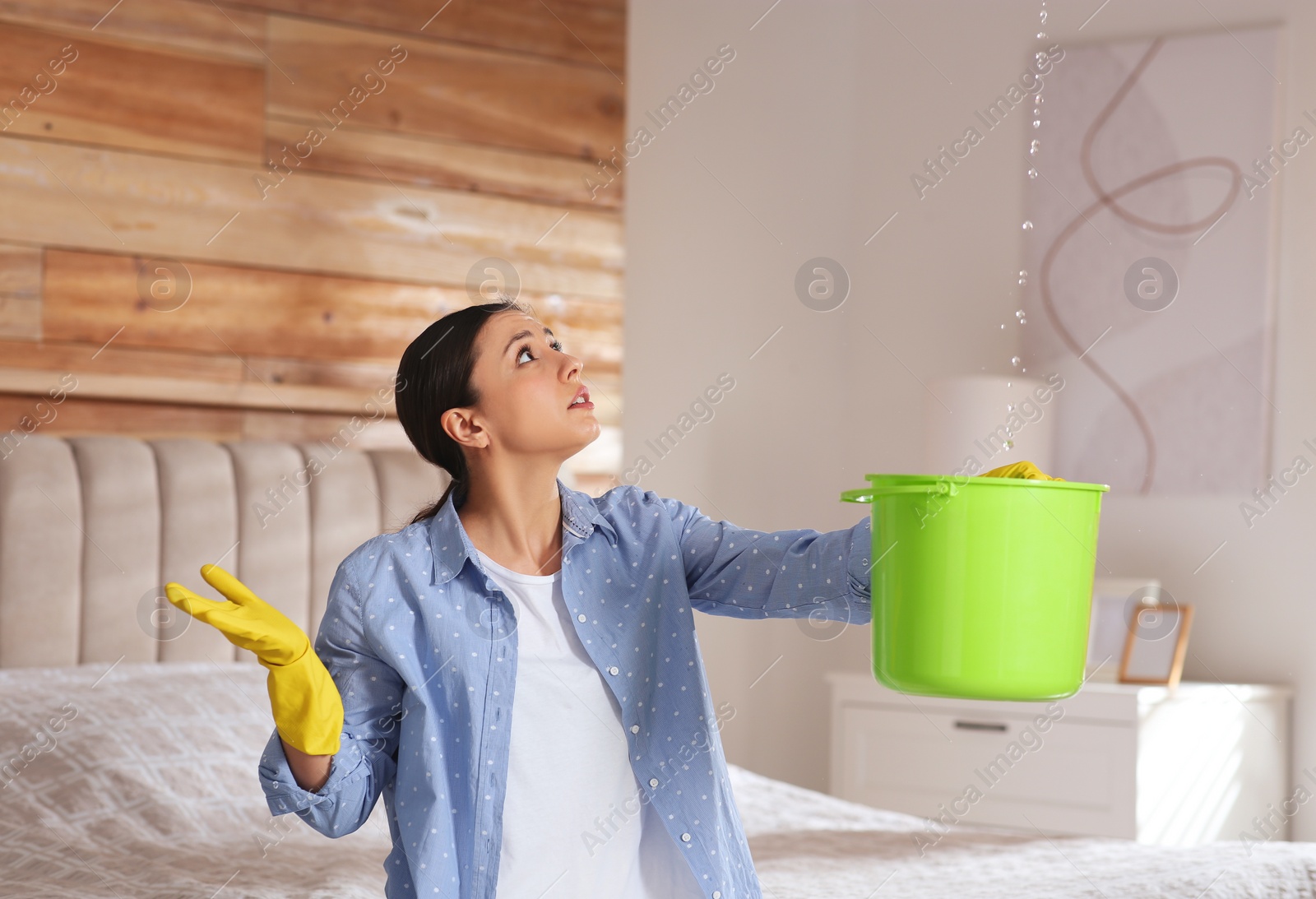 Photo of Young woman collecting leaking water from ceiling in bedroom. Time to call roof repair service