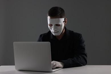 Man in mask and gloves sitting with laptop at white table against grey background