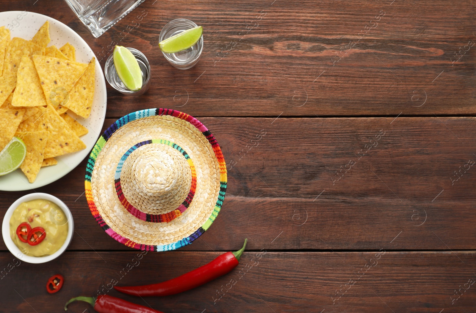 Photo of Mexican sombrero hat, tequila with lime, chili peppers, nachos chips and sauce on wooden table, flat lay. Space for text