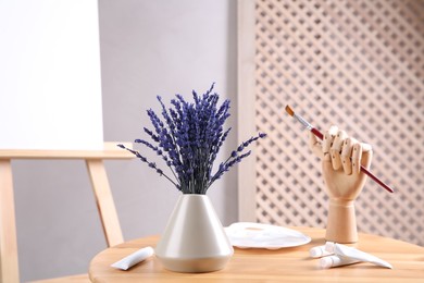 Photo of Bouquet of beautiful preserved lavender flowers, palette, paints and manikin hand with brush on wooden table near easel indoors, closeup