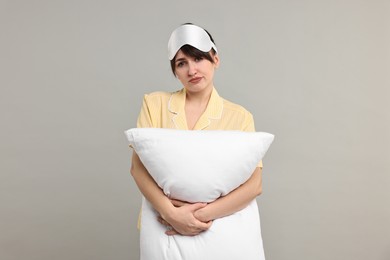 Woman in pyjama and sleep mask holding pillow on grey background