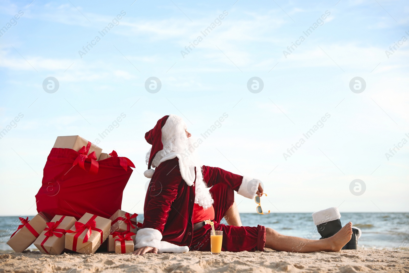 Photo of Santa Claus with bag of presents relaxing on beach, space for text. Christmas vacation