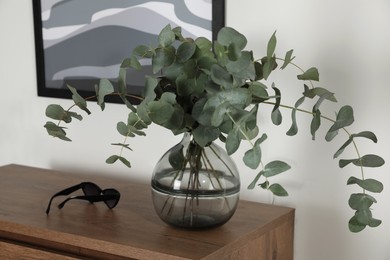 Photo of Sunglasses and glass vase with beautiful eucalyptus branches on wooden table indoors. Interior design