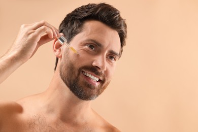 Smiling man applying cosmetic serum onto his face on beige background, closeup. Space for text