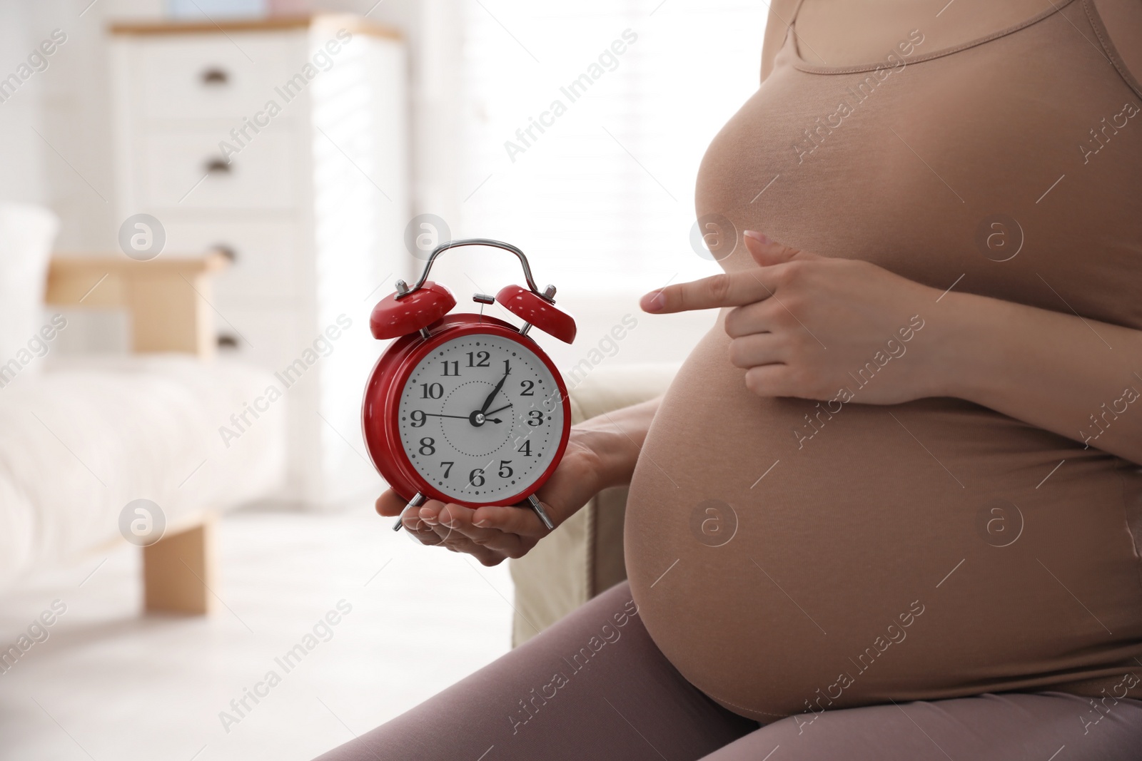 Photo of Young pregnant woman pointing at alarm clock near her belly indoors, closeup. Time to give birth