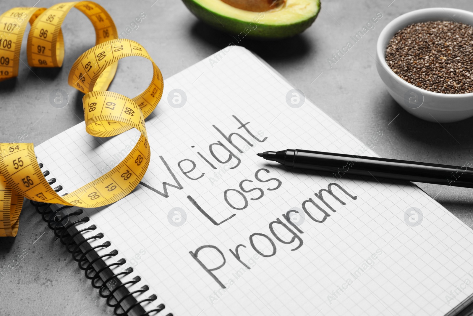 Photo of Notebook with phrase Weight Loss Program, measuring tape, chia seeds and avocado on grey table, closeup