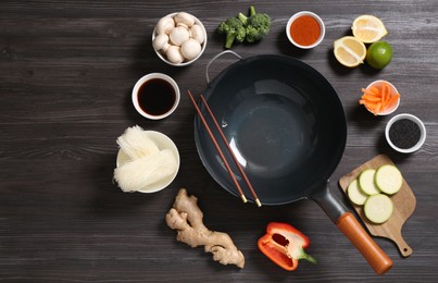 Photo of Empty iron wok and chopsticks surrounded by ingredients on dark grey wooden table, flat lay. Space for text