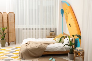 Large comfortable bed, SUP board and green houseplants in stylish bedroom. Interior design
