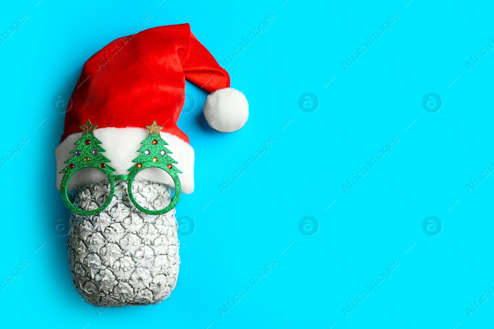 Photo of Top view of pineapple with Santa hat and Christmas glasses on light blue background, space for text. Creative concept