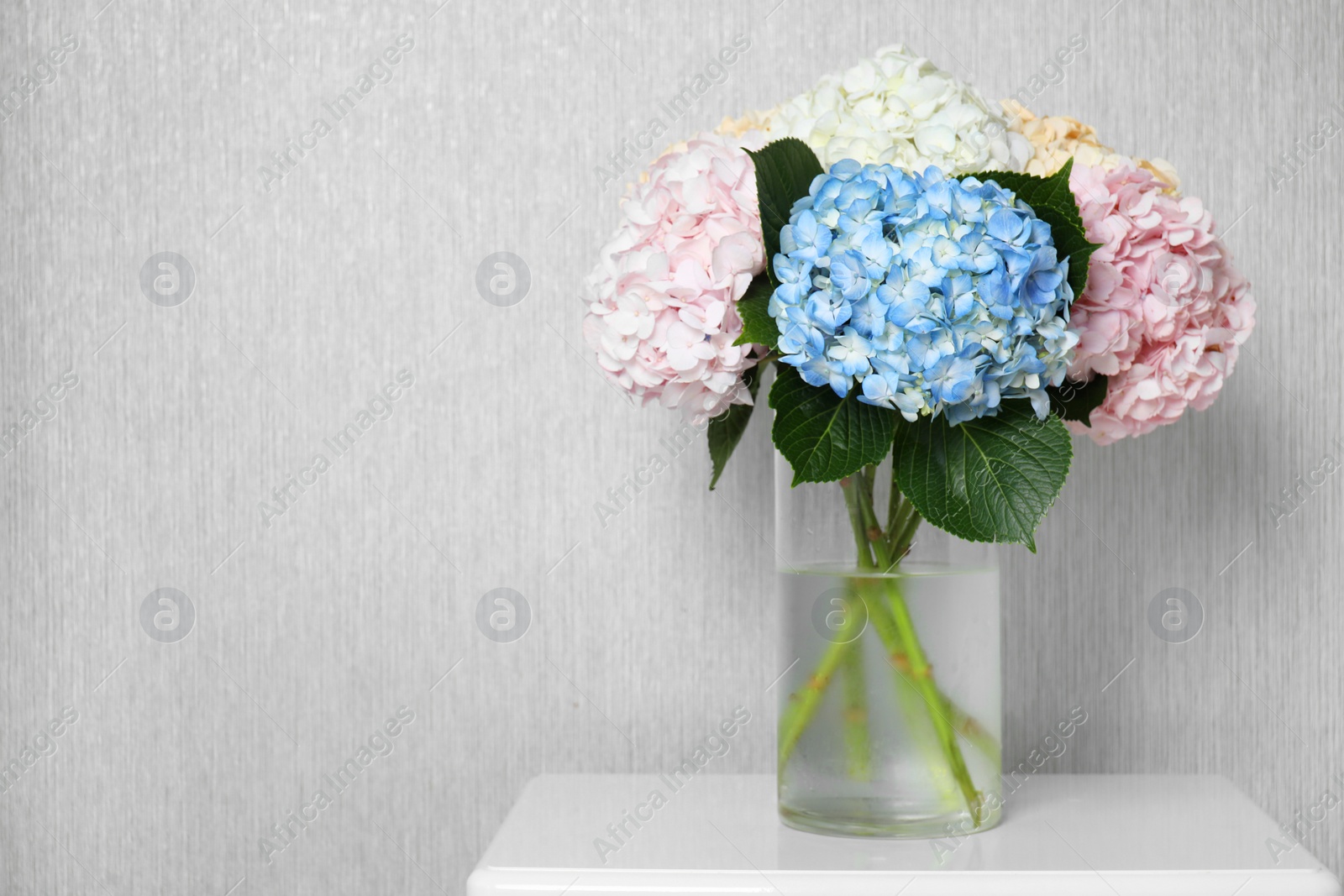 Photo of Beautiful hydrangea flowers in vase on white bedside table near light gray wall. Space for text