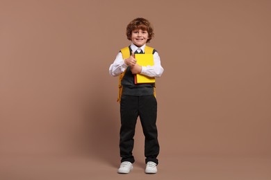 Photo of Happy schoolboy with backpack and books showing thumb up gesture on brown background