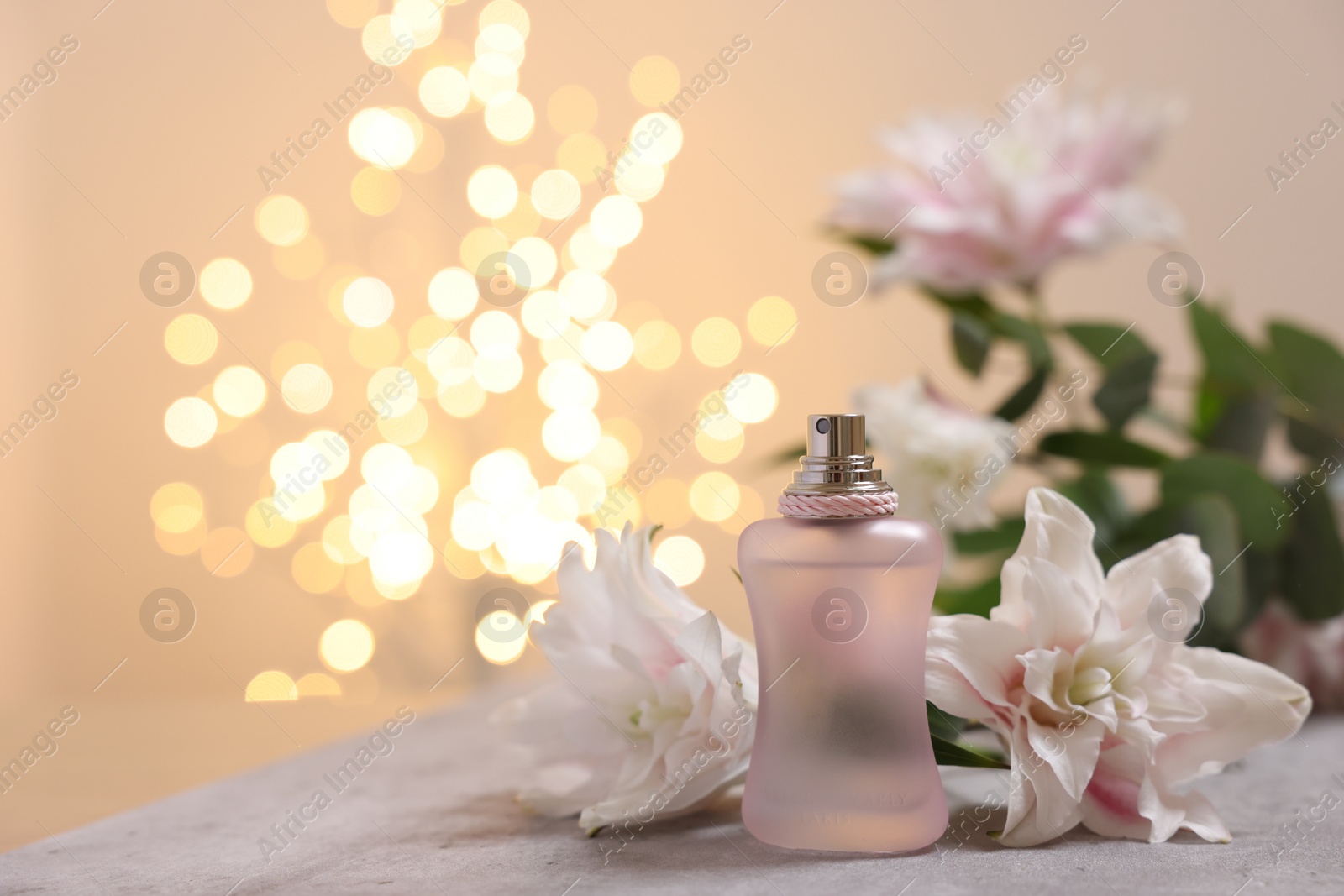 Photo of Bottle of perfume and beautiful lily flowers on table against beige background with blurred lights, closeup. Space for text