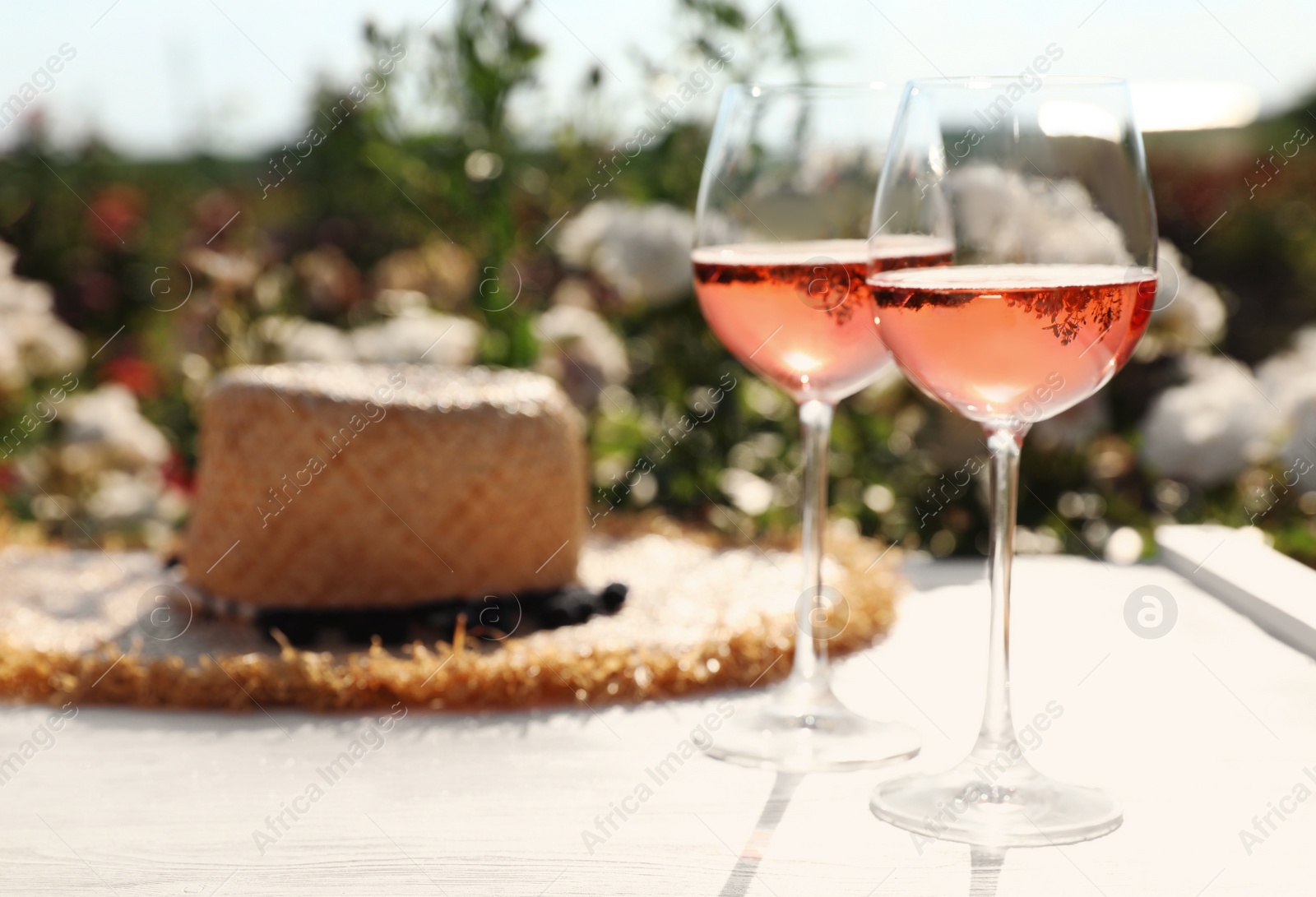 Photo of Glasses of rose wine and straw hat on white wooden table outdoors. Space for text