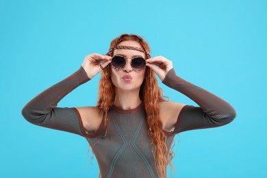 Photo of Stylish young hippie woman in sunglasses sending air kiss on light blue background