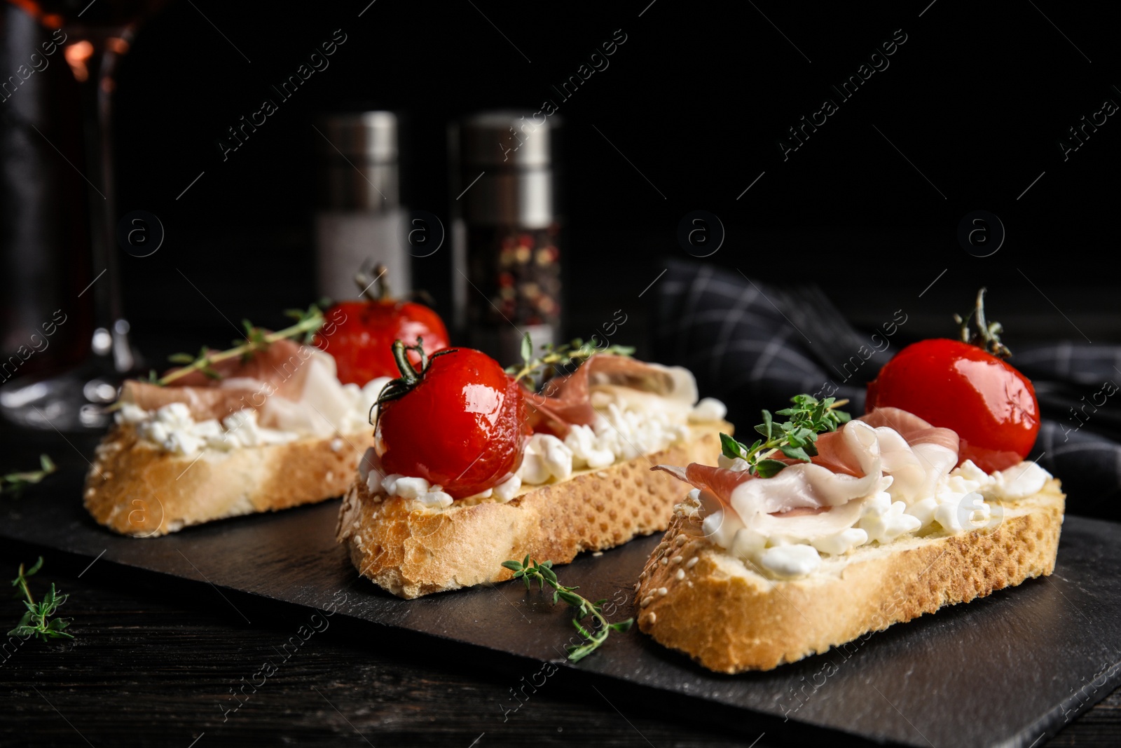 Photo of Slate plate of delicious bruschettas with prosciutto on black wooden table