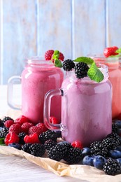 Photo of Mason jars of different berry smoothies and fresh ingredients on white wooden table