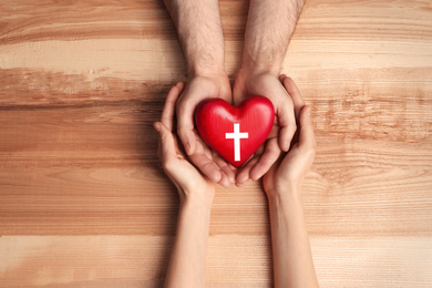 Couple holding heart with cross symbol on wooden background, top view. Christian religion