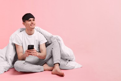Photo of Happy man in pyjama with sleep mask, blanket and smartphone on pink background, space for text