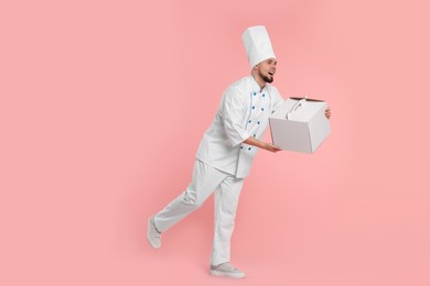 Happy professional confectioner in uniform holding cake box on pink background