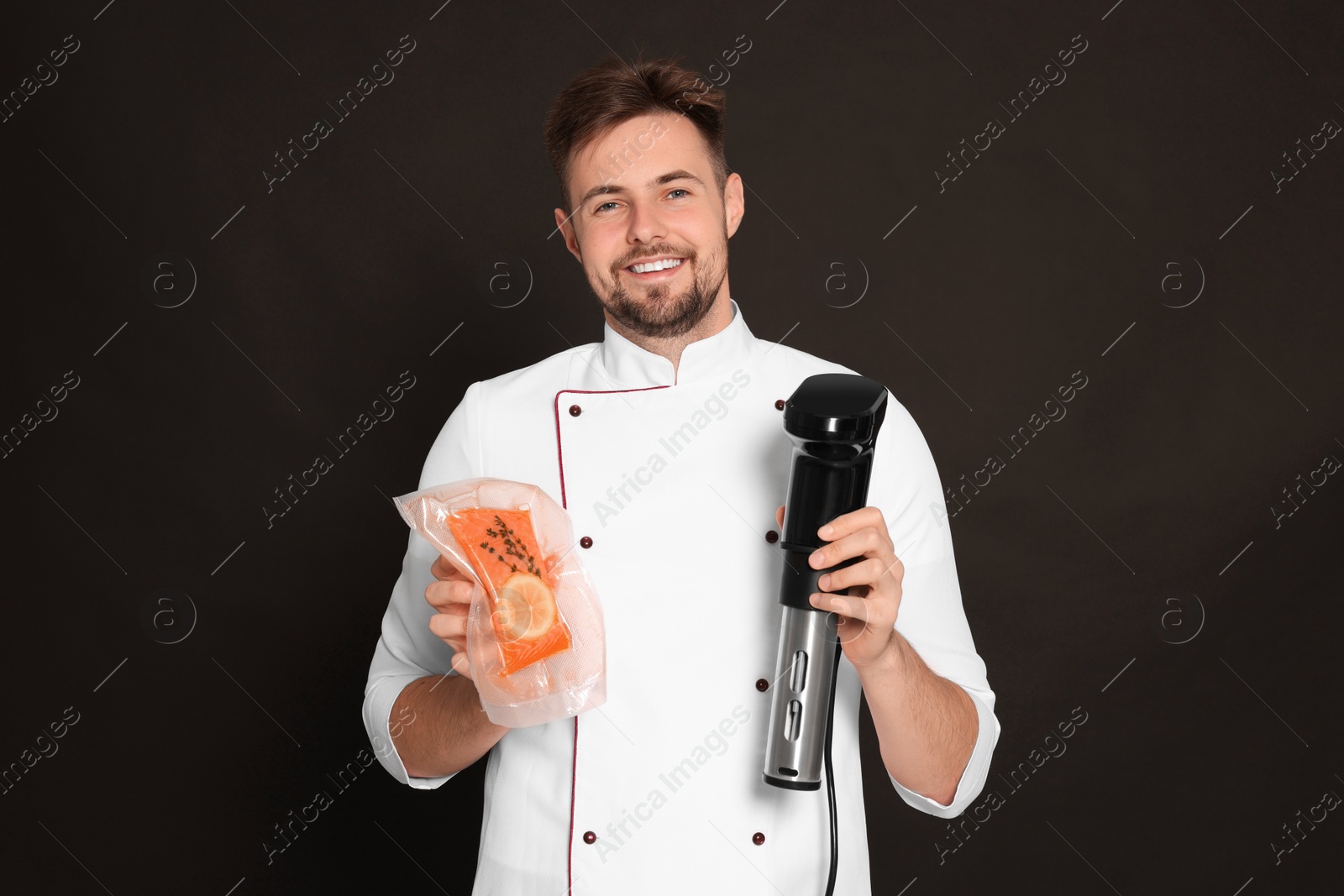 Photo of Smiling chef holding sous vide cooker and salmon in vacuum pack on black background