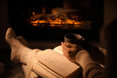 Woman with book and cup near fireplace indoors, closeup. Cozy atmosphere